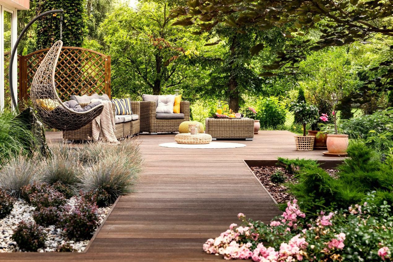 Ein moderner Garten mit Holzterrasse, bequemen Lounge-Möbeln, einem Hängesessel und einem Pavillon, umgeben von üppigem Grün und Blumen.