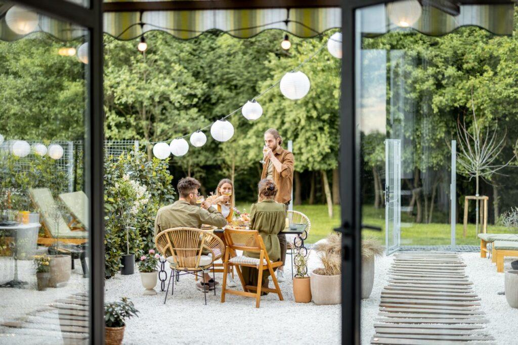 Eine Gruppe von Menschen genießt eine Gartenfeier an einem Holztisch, umgeben von Pflanzen, Lichterketten und einem Pavillon im Hintergrund.