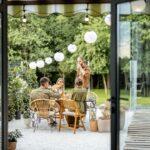 Eine Gruppe von Menschen genießt eine Gartenfeier an einem Holztisch, umgeben von Pflanzen, Lichterketten und einem Pavillon im Hintergrund.
